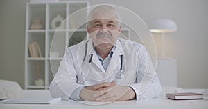 Middle-aged male doctor is greeting participants of webinar or video conference and speaking, portrait in office of
