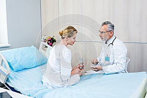 middle aged male doctor with stethoscope over neck pointing at pills to female patient in hospital