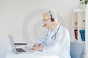 Middle aged male doctor using headset and laptop for online video call consulting of patient. Telemedicine concept for