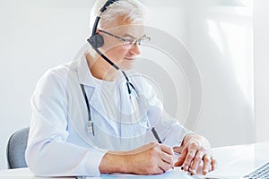 Middle aged male doctor using headset and laptop for online video call consulting of patient. Telemedicine concept for