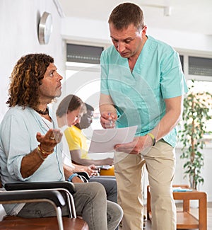 Middle-aged male doctor and visitor sitting on bench discussing medical document