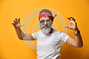 Middle-aged male in sunglasses, red bandana, white t-shirt and bracelet. He scaring you with his hands posing on orange background