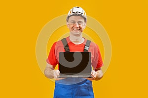 Middle aged man architect engineer wears safety helmet which hand holding laptop with empty screen on yellow studio background.