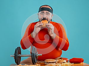 Middle-aged man with beard, dressed in a red turtleneck, headband, posing with burgers and french fries. Blue background