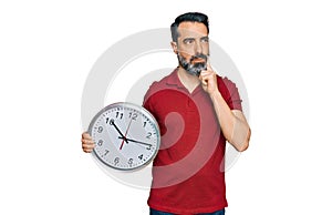 Middle aged man with beard holding big clock serious face thinking about question with hand on chin, thoughtful about confusing