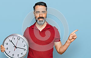 Middle aged man with beard holding big clock smiling happy pointing with hand and finger to the side