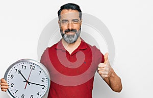 Middle aged man with beard holding big clock smiling happy and positive, thumb up doing excellent and approval sign