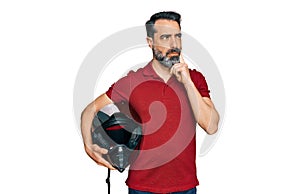 Middle aged man with beard holding motorcycle helmet serious face thinking about question with hand on chin, thoughtful about