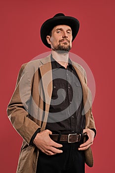 Middle-aged man with beard and mustache, wears black hat and brown jacket posing against a red background. Sincere