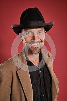 Middle-aged man with beard and mustache, wears black hat and brown jacket posing against a red background. Sincere