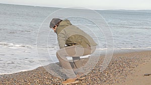 A middle aged man with a beard rolling up his sleeves, squatting down, watering his hands in the tide, wiping his face