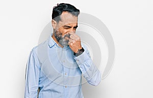 Middle aged man with beard wearing business shirt smelling something stinky and disgusting, intolerable smell, holding breath with