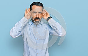 Middle aged man with beard wearing business shirt trying to hear both hands on ear gesture, curious for gossip