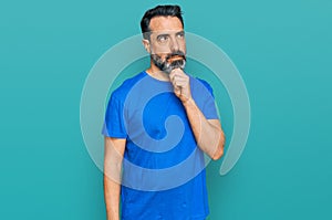 Middle aged man with beard wearing casual blue t shirt with hand on chin thinking about question, pensive expression