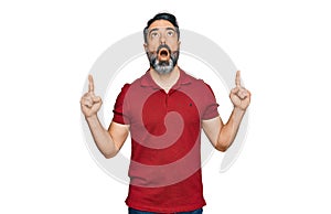 Middle aged man with beard wearing casual red t shirt amazed and surprised looking up and pointing with fingers and raised arms