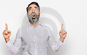 Middle aged man with beard wearing casual white shirt amazed and surprised looking up and pointing with fingers and raised arms