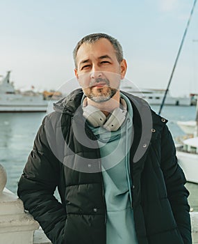 A middle-aged man in a black jacket and full-size headphones in front of yachts.