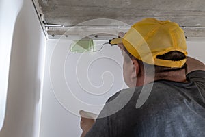 A middle-aged man in dirty clothes and a yellow cap paints a wall with a paint roller. Builder painting a wall in green.