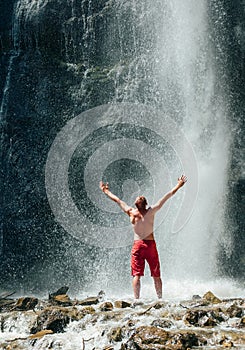 Middle-aged man dressed only red trekking shorts standing under the mountain river waterfall, rose arms up and enjoying the