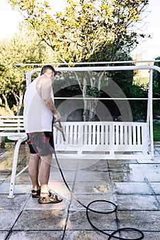 Middle-aged man dressed in summer swimsuit, in the yard of his house cleaning with a pressure water jet machine a white garden
