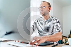 A middle-aged man in eyeglasses typing a text using a modern keyboard computer in his living room. Writing or Distance or