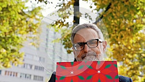Middle-aged man with grey hair and beard sitting on park bench holding red gft bag smiling to camera. Birthday gift