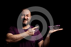 A middle-aged man in his forties holds a toy grocery basket in his hand against a black background and points with a finger to an