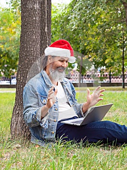 Middle aged man with a laptop in the park. He sitting on the grass in Santa Hat and communicates with relatives on the