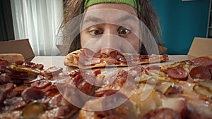 Middle-aged man with long hair and fitness bandage is shown in a close-up shot of a pizza box against a blue wall. Shot