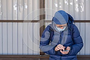 Middle-aged man in medical mask with phone sits in the fall at public transport stop and waits for bus, tram, trolleybus