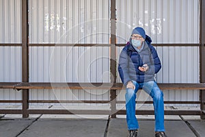 A middle-aged man in a medical mask sits at a public transport stop and waits for a bus. Looks into the distance