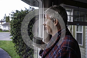 Middle aged man with messy hair and unshaved looking out window with coffee