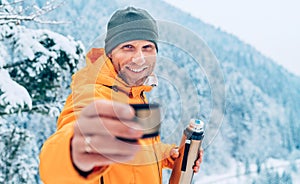 A middle-aged man offering a hot drink cup from a thermos flask dressed in a bright orange softshell jacket while he trekking the