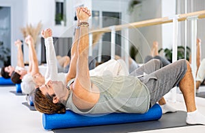 Middle-aged man practicing pilates with roller on gray mat in gym room