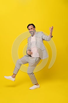 Middle aged man pretend playing guitar laughing wearing white suit isolated on yellow background. Handsome mature