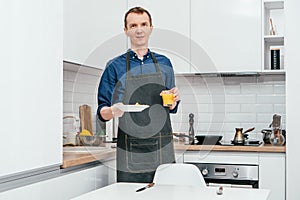 Middle-aged man with short ginger hair in blue shirt, black apron stand holding glass of orange juice and white plate.