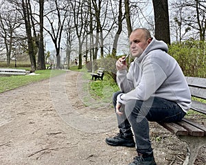 A middle-aged man sits on a park bench in the spring and smokes