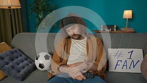 A middle aged man is sitting on a sofa in a room. Next to him is a soccer ball and a tablet. He fell asleep holding a TV