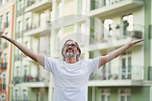 Middle aged man spread his hands in the air praising God standing outdoors wearing white t-shirt and eye glasses. Middle