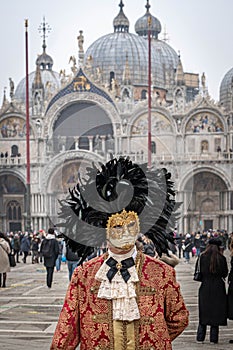 A middle-aged man in a vintage costume with a lace collar, wearing a carnival mask bordered by a circle of high black feathers