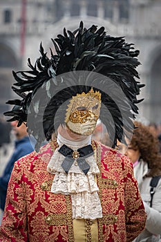 A middle-aged man in a vintage costume with a lace collar, wearing a carnival mask bordered by a circle of high black feathers