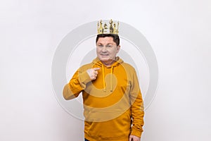 Middle aged man wearing golden crown and pointing himself, looking at camera with smile.