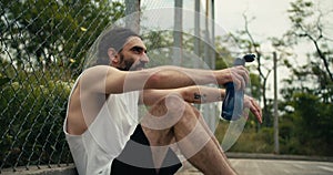 A middle-aged man in a white T-shirt sits on a basketball court near the fence, rests and drinks water from a special