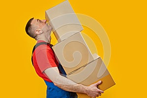 Middle-aged man in workers uniform holds large heavy boxes. Portrait of loader in studio on yellow background. Side view. Copy