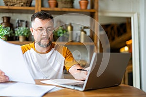 A middle-aged man works with papers at home in front of a laptop monitor. He is sitting with glasses and typing text.