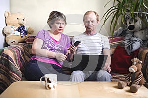 A middle-aged married couple siting at home with a laptop. On the sofa plush toy