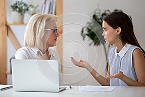 Middle-aged and millennial female colleagues arguing seated at workplace photo