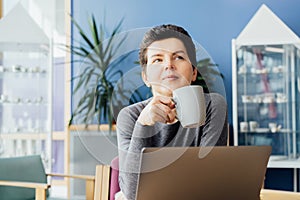 Middle aged minded pensive employee business woman in casual clothes holds coffee cup works at office desk with pc laptop.