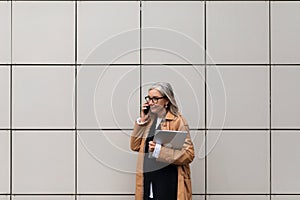 Middle aged older business woman with a laptop in her hands speaks on a mobile phone against a beige ceramic tile wall