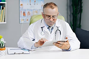 Middle aged older male doctor in white uniform holding digital computer tablet in hands, managing patients visits